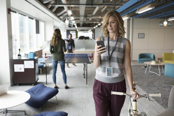 Businesswoman with smart phone walking with bicycle in office while colleagues play ping pong in background
