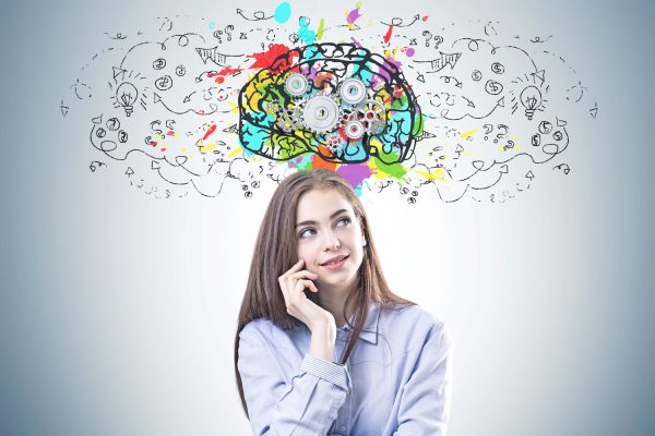 Dreamy young woman with brown hair wearing a blue shirt and business suit pants is looking upwards and thinking. A gray wall background with a cog brain and arrows on it.